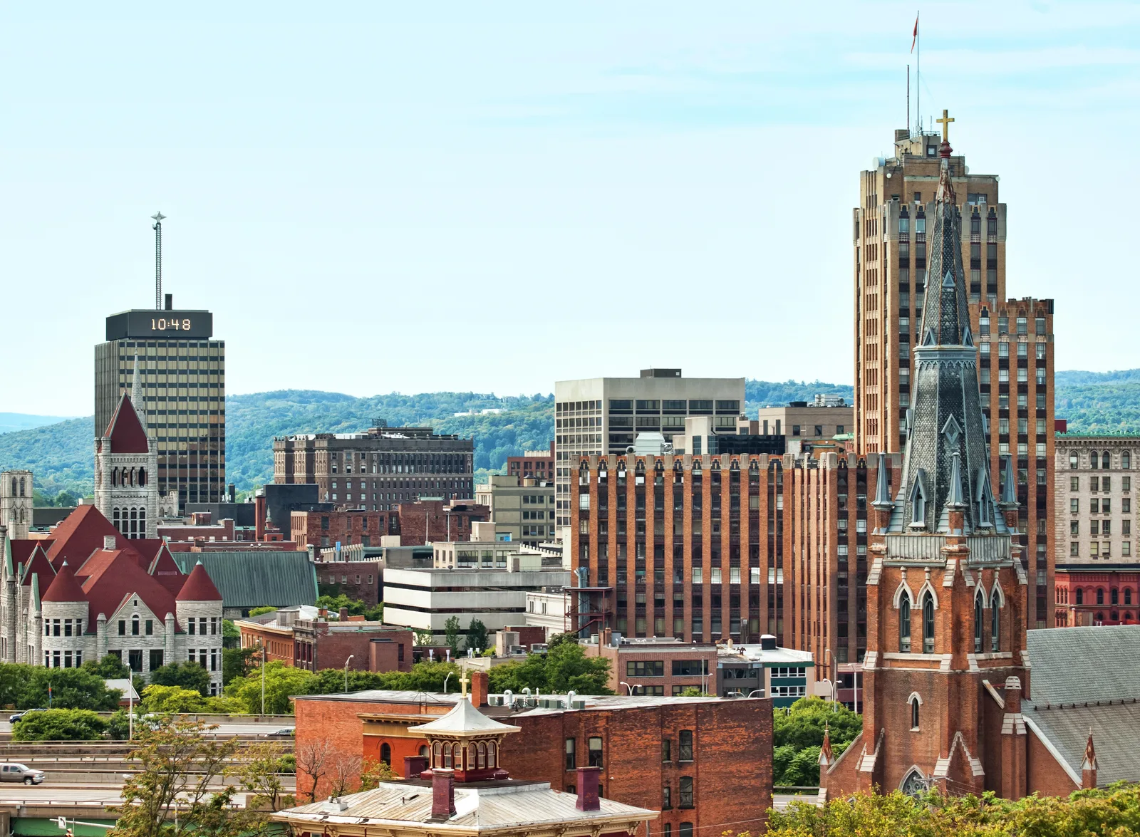 Downtown skyline with historic churches, brick buildings, modern towers, and distant green hills under a bright sky.