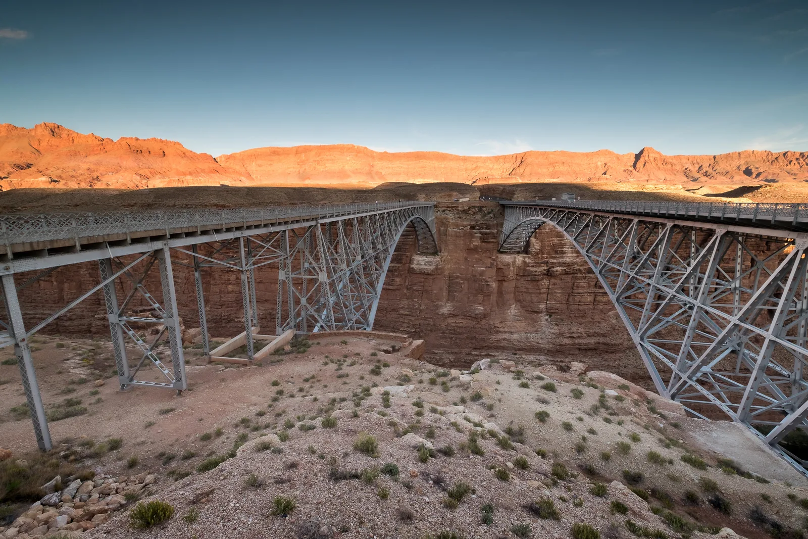 Two steel truss highway bridges span a deep, rocky canyon under a clear sky.