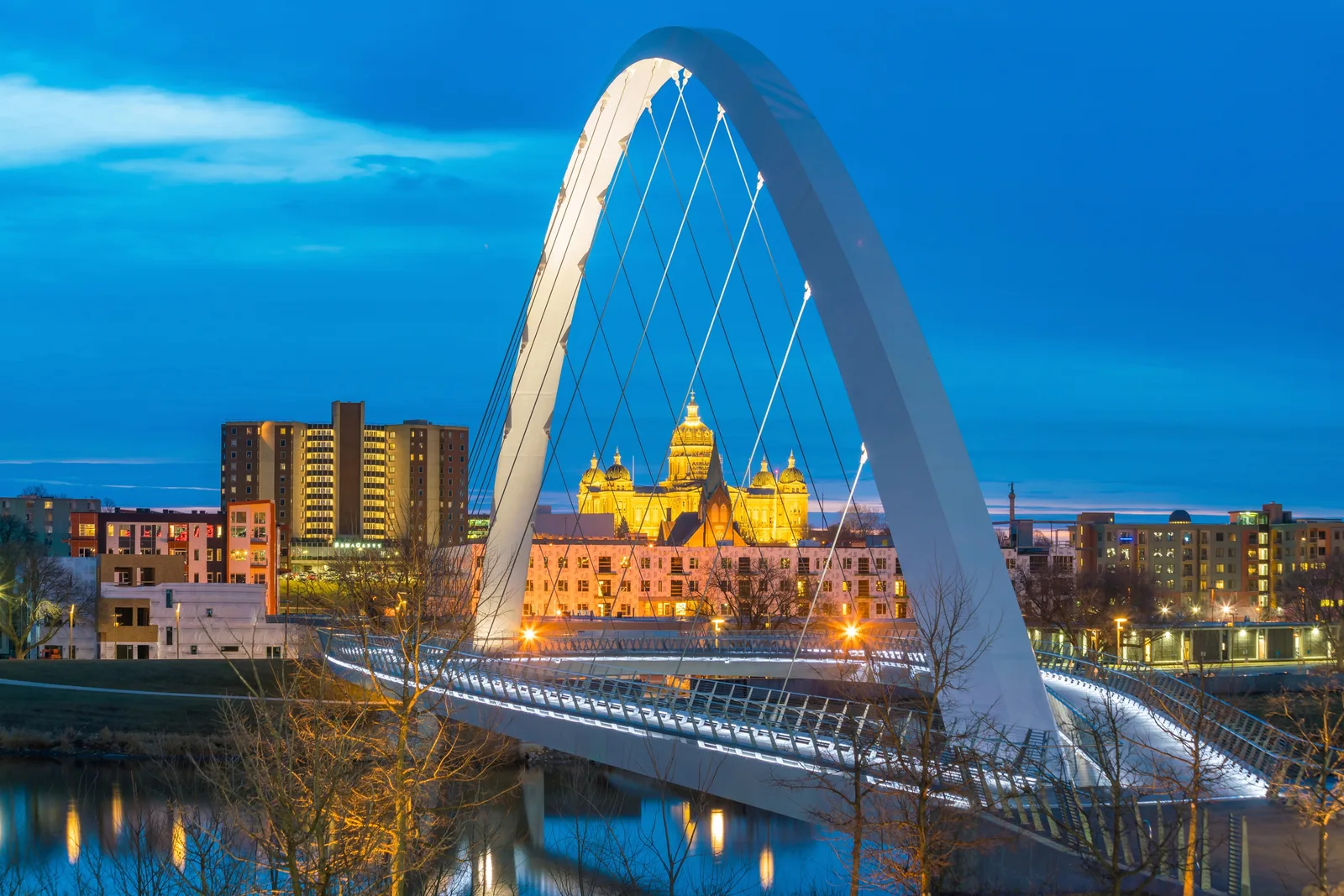An illuminated arched pedestrian bridge framing the city skyline in Des Moines at dusk.