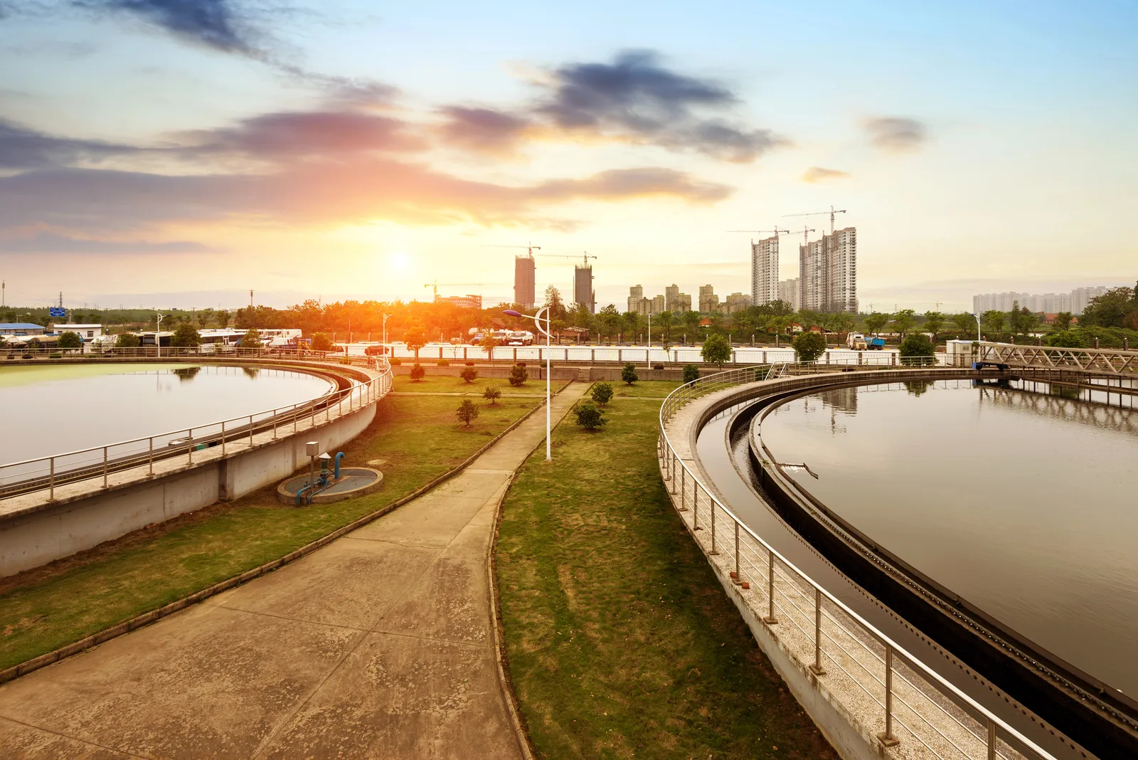 Two large circular wastewater treatment tanks in foreground with grass and walkways; city skyline in background at sunset.