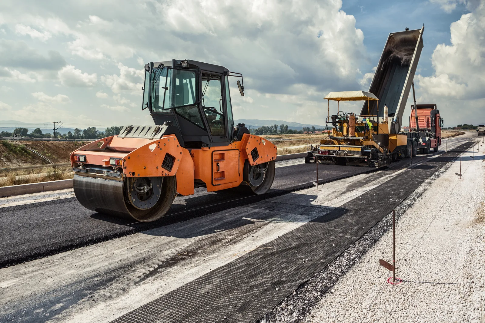 Orange road roller and yellow asphalt paver lay new asphalt on a road construction site under a cloudy sky.